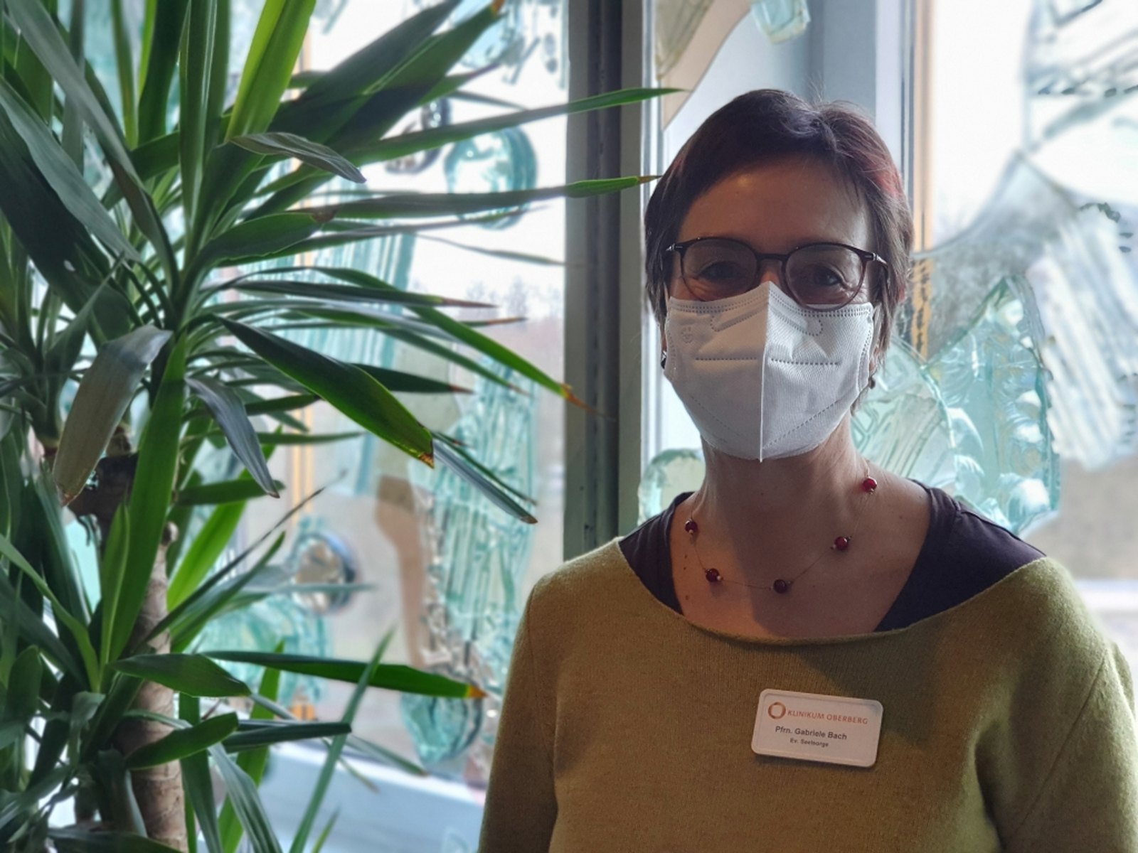 Das Bild zeigt Gabriele Bach mit FFP2-Maske vor dem bunten Fenster in der Krankenhauskapelle   