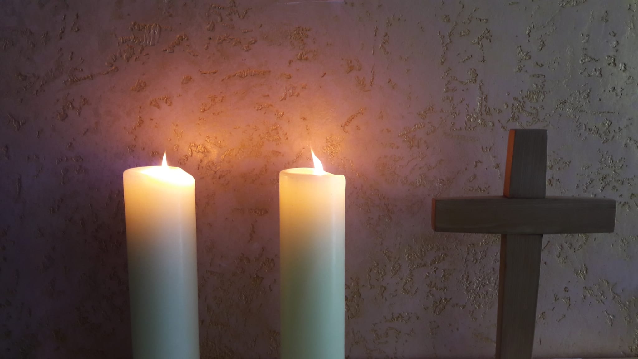 Das Bild zeigt zwei große weiße Kerzen neben einem Kreuz aus Holz z 